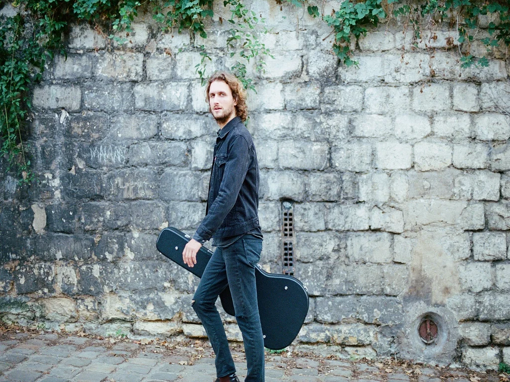 Greg Novan est debout, de profil, regarde l'objectif et est habillé en noir et porte sa guitare en étui noir, un fond de mur en pierres en arrière plan