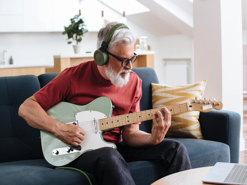 un homme blanc aux cheveux blancs porte un tshirt rouge, est assis sur un canapé gris et joue de la guitare électrique. Il porte un casque sur les oreilles.