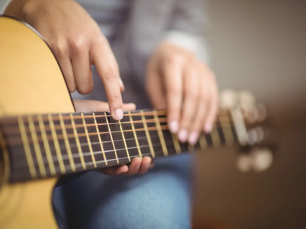 L'image montre un manche de guitare sèche sur lequel se trouvent 3 mains. Une qui soutient le manche et les deux autres qui se trouvent sur les cordes