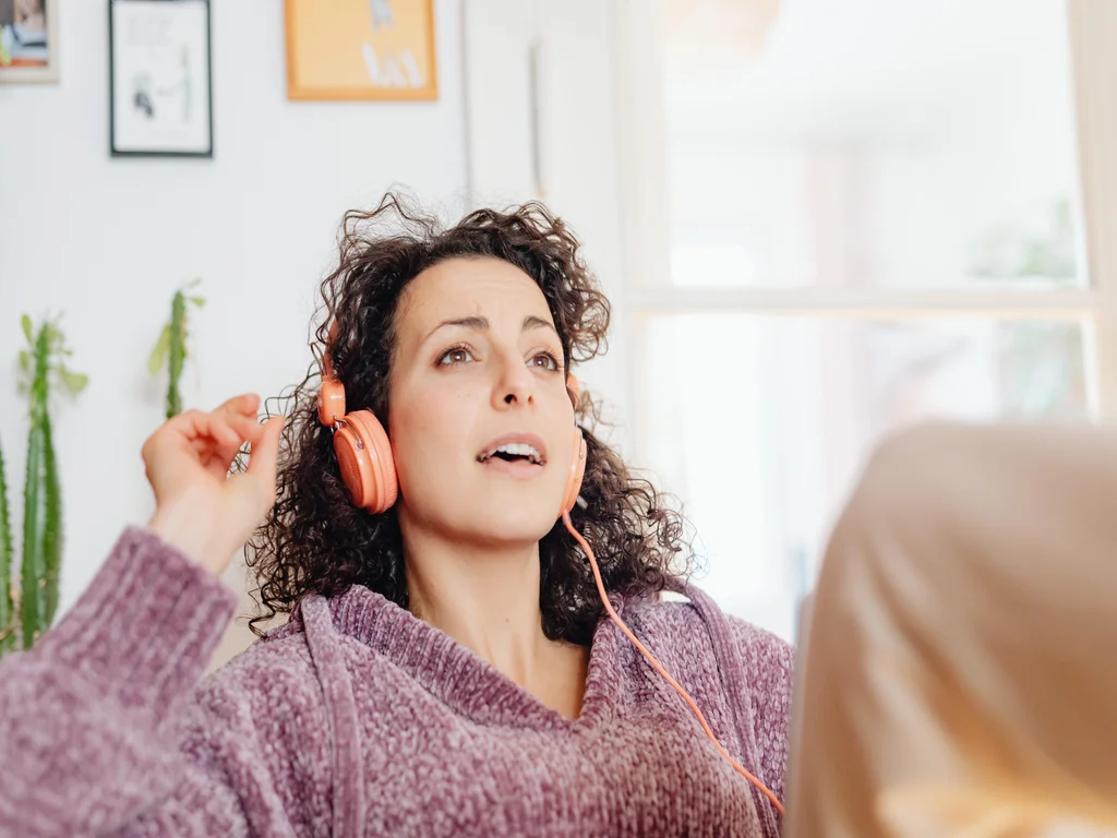 une femme blanche, brune, aux cheveux mi-longs est assise et porte un casque de musique sur les oreilles. Le casque est orange et la femme a la bouche ouverte comme si elle s'entrainait aux différentes techniques de chant, elle porte un pull de couleur mauve. 
